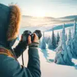 A photographer dressed in warm clothing taking pictures in a snow-covered landscape with snow-covered trees and a clear sky, capturing the serene winter scenery.