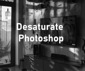 A black and white photo of a room with sunlight streaming through a window, casting shadows on the wall. The room contains a television and speakers on a cabinet, with framed items on the wall. Text overlay reads “Desaturate Photoshop” along with the Photoshop logo.